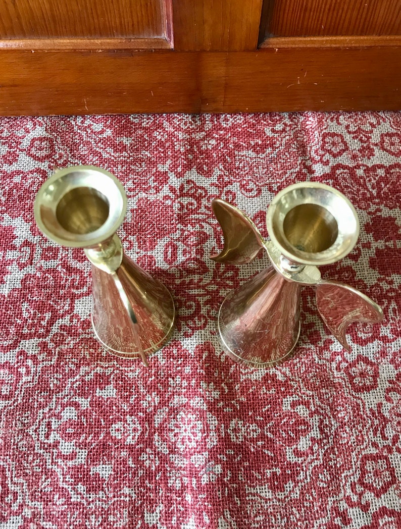 May include: Two gold metal candle holders with a wing design. The candle holders are on a red and white patterned surface.