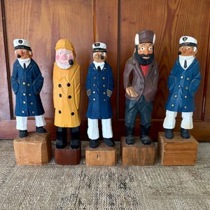 May include: Five hand-carved wooden sailor figurines. The sailors are dressed in blue coats, yellow raincoats, and brown jackets. Each figurine stands on a small wooden block. The sailor's hats are white.