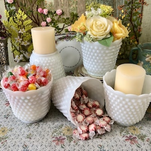 May include: A collection of white hobnail glass containers filled with wrapped candies and a candle. The containers include a vase, a small bowl, and a cone-shaped holder. Yellow roses and a candle are also present.