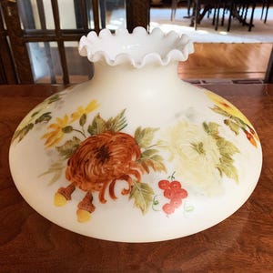 May include: Off-white glass lampshade with a ruffled edge, hand-painted with colorful floral designs. The flowers include orange, yellow, and white chrysanthemums, with green leaves and red berries. A decorative lighting element.