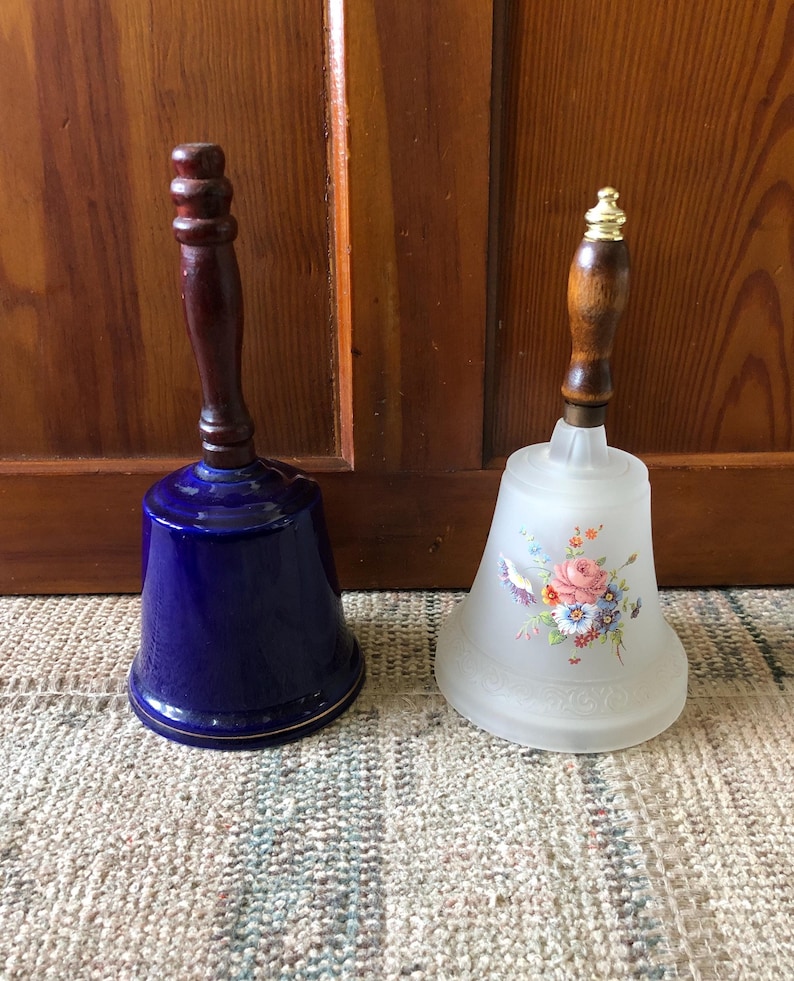 May include: Two vintage decorative bells with wooden handles. One bell is blue with a gold trim and the other is white with a floral design.