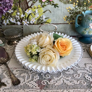 Puede incluir: Bandeja de servir de vidrio blanco lechoso con un asa plateada y bordes festoneados. La bandeja est&aacute; decorada con una banda plateada alrededor del borde. La bandeja est&aacute; llena de flores artificiales, incluyendo rosas y aliento de beb&eacute;.