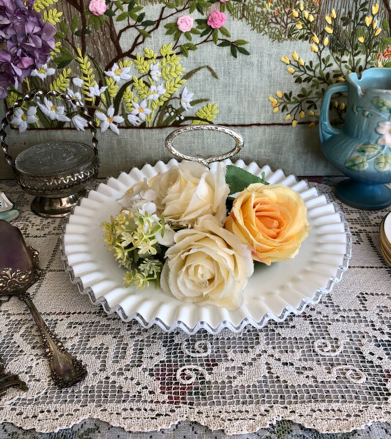 Puede incluir: Bandeja de servir de vidrio blanco lechoso con un asa plateada y bordes festoneados. La bandeja est&aacute; decorada con una banda plateada alrededor del borde. La bandeja est&aacute; llena de flores artificiales, incluyendo rosas y aliento de beb&eacute;.