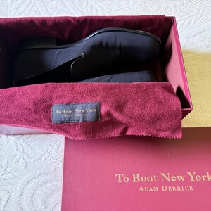 Puede incluir: Zapatos de vestir negros dentro de una caja burdeos con un pa&ntilde;o burdeos. La caja tiene el texto "To Boot New York" y "Adam Derrick" impreso. Los zapatos son de un material oscuro y liso.