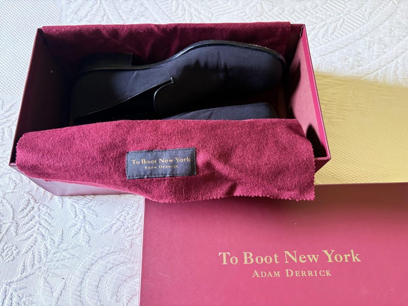 Puede incluir: Zapatos de vestir negros dentro de una caja burdeos con un pa&ntilde;o burdeos. La caja tiene el texto "To Boot New York" y "Adam Derrick" impreso. Los zapatos son de un material oscuro y liso.