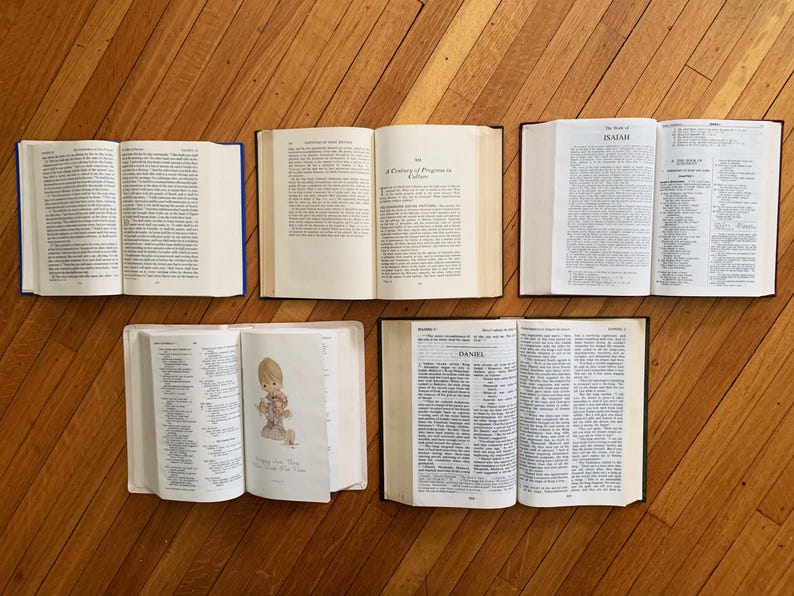 Op de afbeelding: Een luchtfoto van vijf open boeken op een houten oppervlak. De boeken hebben cr&egrave;mekleurige pagina's en zijn in een halve cirkel gerangschikt. De boekbanden zijn in verschillende kleuren, waaronder blauw en zwart. De boeken lijken religieuze teksten te zijn.
