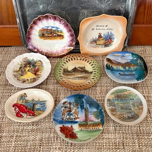 May include: A collection of eight decorative ceramic souvenir plates featuring various illustrations including a lobster, a railway station, a mountain lake, and Canadian landmarks with text such as St. Catharines, Canada and Seldom Seen Valley Mine.