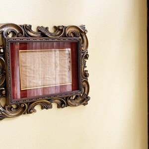 May include: A gold ornate picture frame with a red mat and a blank white paper insert.