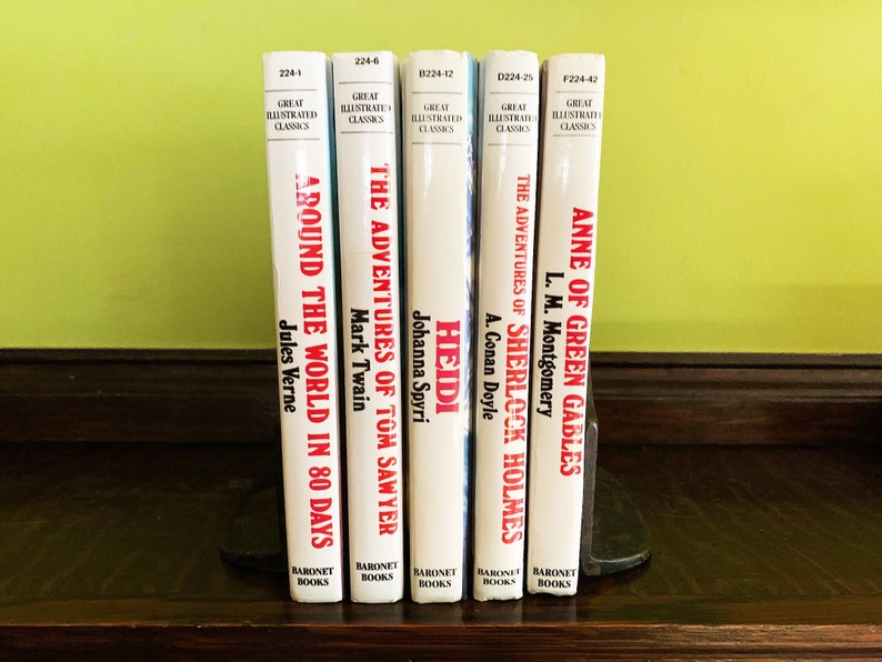 May include: A set of five classic literature books with white covers and red titles. The books are arranged vertically on a wooden shelf. The titles are "Around the World in 80 Days", "The Adventures of Tom Sawyer", "Heidi", "The Adventures of Sherlock Holmes", and "Anne of Green Gables".