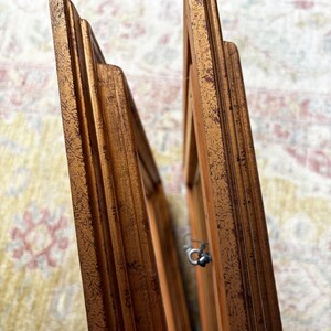 May include: Two rectangular picture frames with a distressed gold finish and a stepped, tiered profile. The frames are positioned at an angle, revealing the side edges and a small metal hanging hook attached to the inner side of one frame.