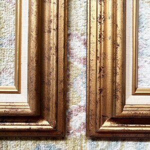May include: Two rectangular picture frames featuring a metallic gold finish with dark brown distressed detailing. Each frame includes a light beige fabric matting border surrounding the inner opening, showcasing a multi-layered profile design.