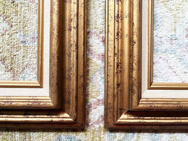 May include: Two rectangular picture frames featuring a metallic gold finish with dark brown distressed detailing. Each frame includes a light beige fabric matting border surrounding the inner opening, showcasing a multi-layered profile design.