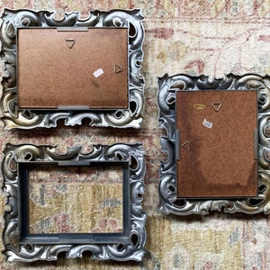 May include: Three ornate silver picture frames with brown cardboard backing. The frames have a decorative, ornate design with a silver finish.