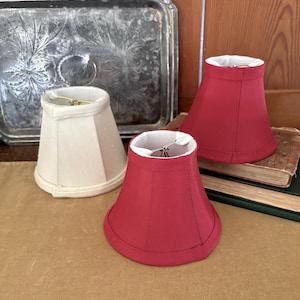 Op de afbeelding: Drie kleine lampenkappen. Twee zijn felrood en &eacute;&eacute;n is gebroken wit. Elke kap heeft een witte binnenkant en een goudkleurig draadframe. De kappen zijn gerangschikt op een bruin oppervlak, met een zilveren dienblad en boeken op de achtergrond.