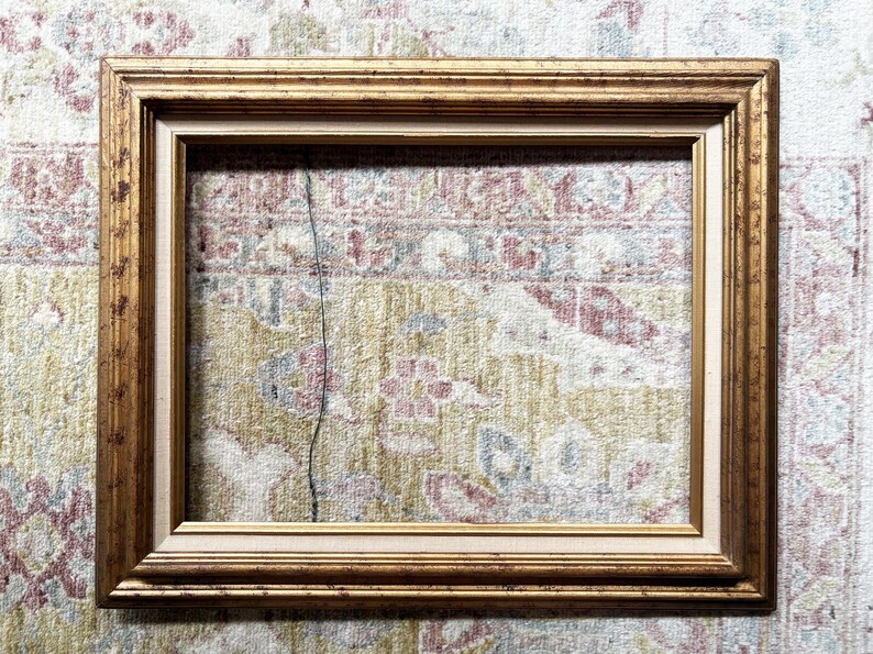 May include: A rectangular picture frame featuring a distressed gold finish with a multi-tiered profile. The frame includes an inner border with a light beige fabric-like texture, creating a layered appearance for displaying artwork or photographs.