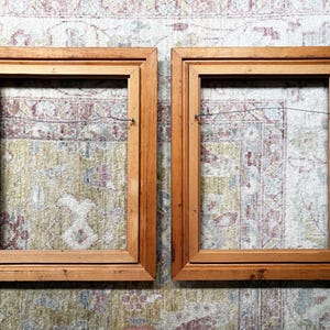 May include: Two rectangular wooden picture frames with a natural light brown finish. Each frame features a stepped inner edge profile and a thin metal hanging wire attached to the back side, spanning horizontally across the open centre area.