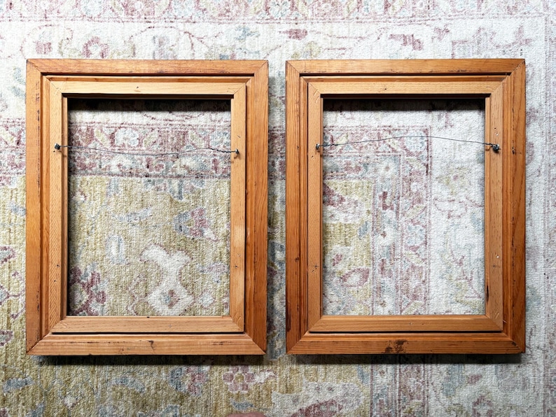 May include: Two rectangular wooden picture frames with a natural light brown finish. Each frame features a stepped inner edge profile and a thin metal hanging wire attached to the back side, spanning horizontally across the open centre area.
