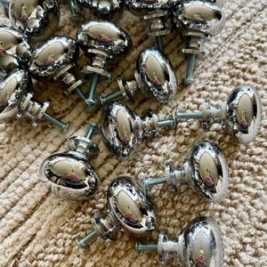 May include: A group of 18 silver-toned cabinet knobs with a rounded oval shape. The knobs have a shiny, reflective finish and are arranged in a random pattern on a light brown, textured surface.