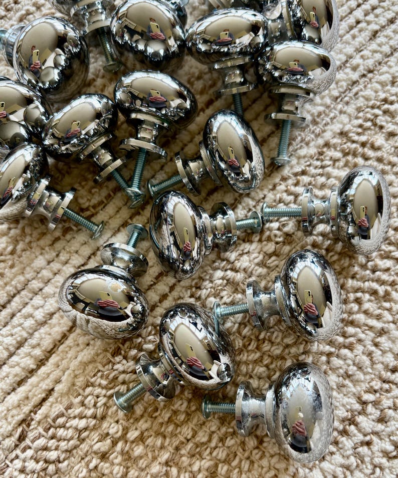 May include: A group of 18 silver-toned cabinet knobs with a rounded oval shape. The knobs have a shiny, reflective finish and are arranged in a random pattern on a light brown, textured surface.