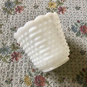 May include: A white hobnail glass vase with a scalloped rim. The vase has a textured surface with small, raised bumps and vertical ridges. The vase is empty and sits on a floral patterned fabric.