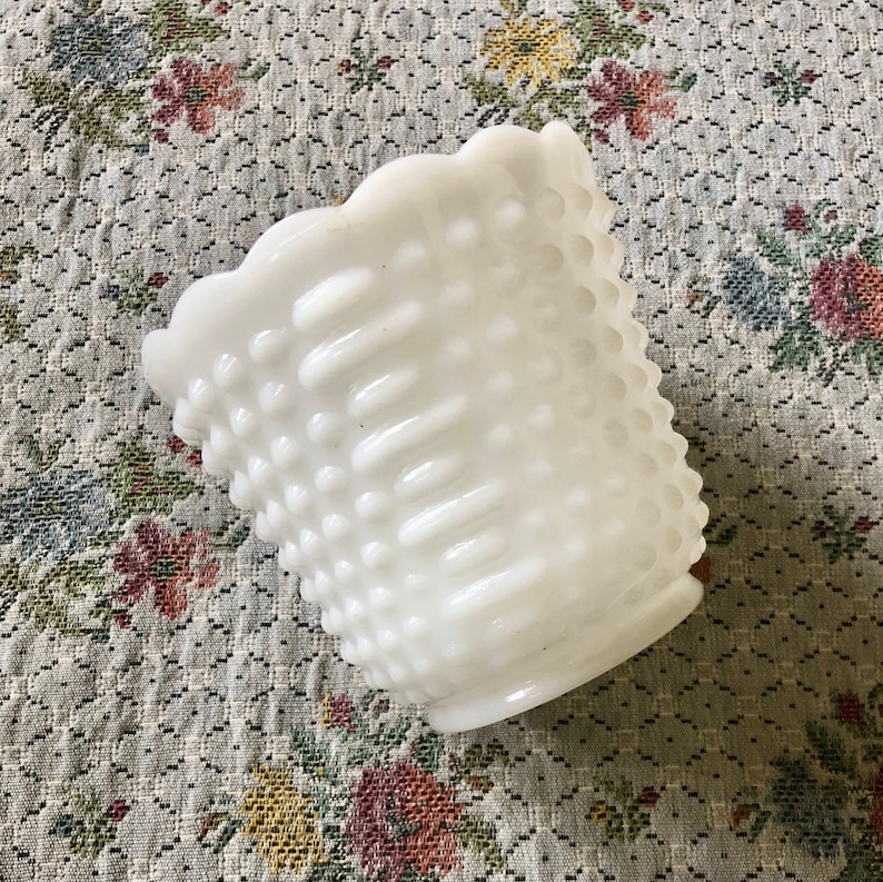 May include: A white hobnail glass vase with a scalloped rim. The vase has a textured surface with small, raised bumps and vertical ridges. The vase is empty and sits on a floral patterned fabric.