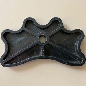 K&ouml;nnte beinhalten: Ein schwarzes Kunststoffobjekt mit einer einzigartigen Form, das ein zentrales Loch und strahlenf&ouml;rmige St&uuml;tzbalken aufweist. Das Objekt hat einen wellenf&ouml;rmigen, konturierten Rand und scheint ein Bestandteil eines gr&ouml;&szlig;eren Artikels zu sein. Die Oberfl&auml;che hat ein leicht strukturiertes Aussehen.