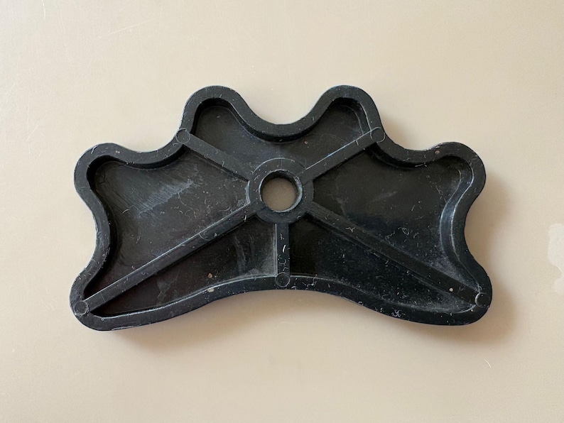 K&ouml;nnte beinhalten: Ein schwarzes Kunststoffobjekt mit einer einzigartigen Form, das ein zentrales Loch und strahlenf&ouml;rmige St&uuml;tzbalken aufweist. Das Objekt hat einen wellenf&ouml;rmigen, konturierten Rand und scheint ein Bestandteil eines gr&ouml;&szlig;eren Artikels zu sein. Die Oberfl&auml;che hat ein leicht strukturiertes Aussehen.