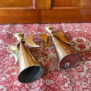 May include: Two gold angel candle holders with wings and a cone-shaped base. The candle holders are on a red and white patterned surface.