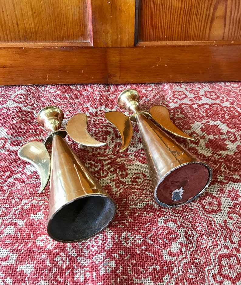 May include: Two gold angel candle holders with wings and a cone-shaped base. The candle holders are on a red and white patterned surface.