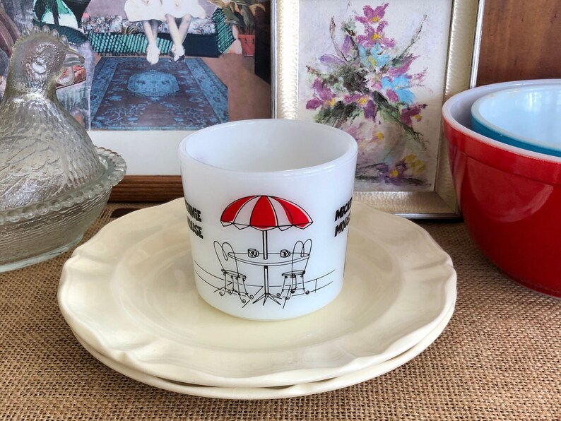May include: White ceramic mug with a black and red illustration of a red and white striped umbrella, two chairs, and a table. The mug says "Nostalgia Village" on the side.