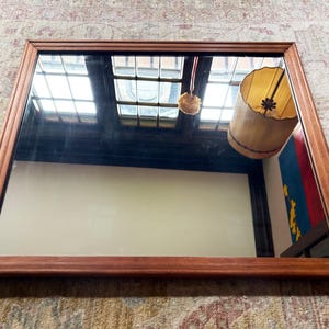 May include: Rectangular wooden-framed mirror. The frame is a warm brown color. The mirror reflects a room with a window, a hanging lamp, and a colorful wall hanging. The mirror is resting on a patterned rug.