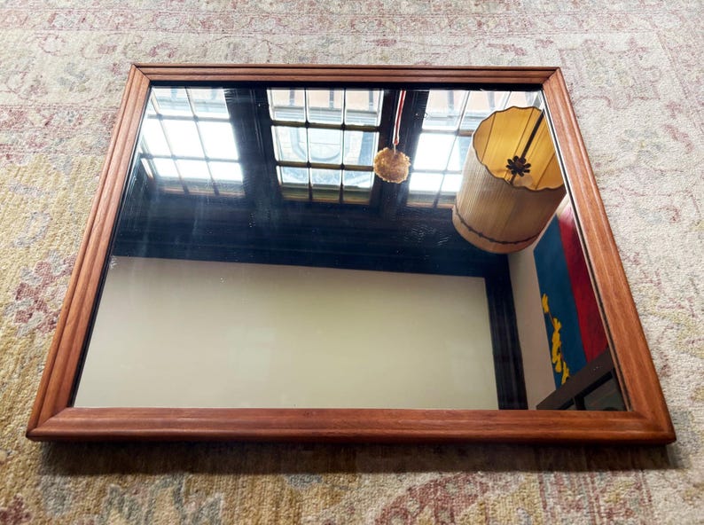 May include: Rectangular wooden-framed mirror. The frame is a warm brown color. The mirror reflects a room with a window, a hanging lamp, and a colorful wall hanging. The mirror is resting on a patterned rug.