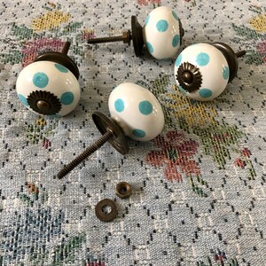 Puede incluir: Cuatro pomos de armario de cer&aacute;mica blanca con lunares azules y detalles en lat&oacute;n. Los pomos tienen un dise&ntilde;o de tornillo y se muestran sobre una tela con estampado floral.