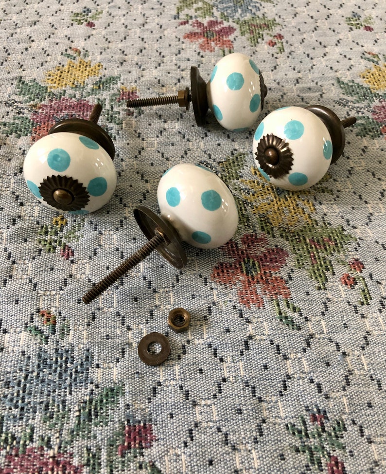 Puede incluir: Cuatro pomos de armario de cer&aacute;mica blanca con lunares azules y detalles en lat&oacute;n. Los pomos tienen un dise&ntilde;o de tornillo y se muestran sobre una tela con estampado floral.