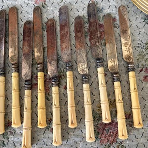 Pode incluir: Um conjunto de facas antigas com l&acirc;minas de prata e cabos em estilo bambu. As l&acirc;minas apresentam padr&otilde;es florais gravados e sinais de desgaste. Os cabos s&atilde;o de cor creme. As facas est&atilde;o dispostas sobre uma toalha de mesa com padr&atilde;o floral.