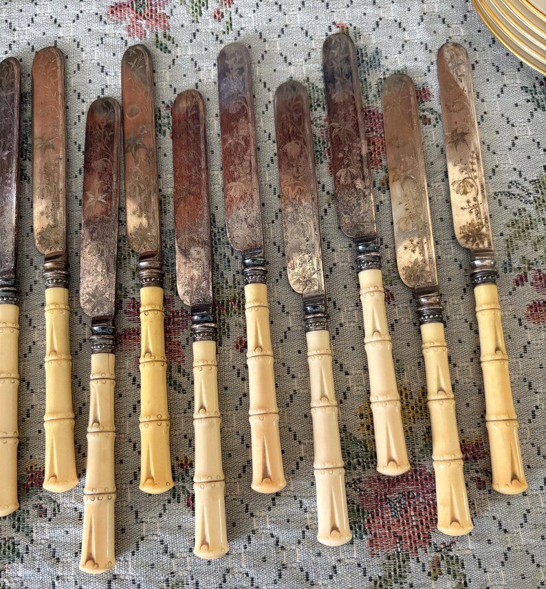 Pode incluir: Um conjunto de facas antigas com l&acirc;minas de prata e cabos em estilo bambu. As l&acirc;minas apresentam padr&otilde;es florais gravados e sinais de desgaste. Os cabos s&atilde;o de cor creme. As facas est&atilde;o dispostas sobre uma toalha de mesa com padr&atilde;o floral.