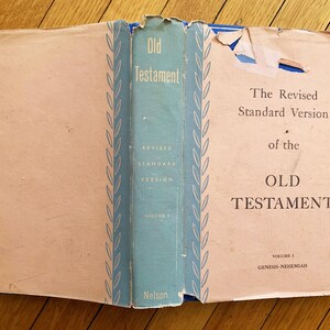 Op de afbeelding: Open boek met de titel "Oude Testament" met een lichtblauwe rug. De tekst "The Revised Standard Version" is zichtbaar. Het boek ligt open op een houten oppervlak.