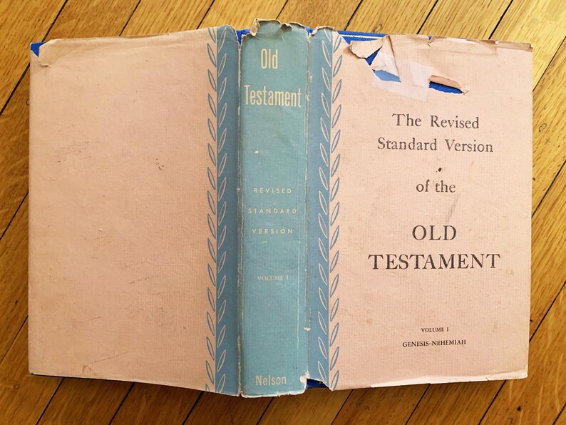 Op de afbeelding: Open boek met de titel "Oude Testament" met een lichtblauwe rug. De tekst "The Revised Standard Version" is zichtbaar. Het boek ligt open op een houten oppervlak.