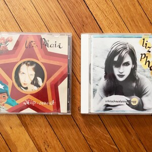 May include: Two Liz Phair music CDs. The first CD is red, white, and blue with a star and a picture of a person. The title is "Whip-Smart". The second CD is white with a black and white photo of Liz Phair. The title is "Whitechocolatespaceegg".