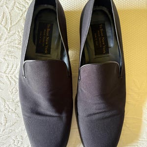 Puede incluir: Un par de zapatos sin cordones de color gris oscuro. Los zapatos tienen una puntera redondeada y un acabado liso y mate. El interior de cada zapato tiene una etiqueta que dice "To Boot New York Adam Derrick MADE IN ITALY". Los zapatos se muestran sobre una superficie blanca estampada.