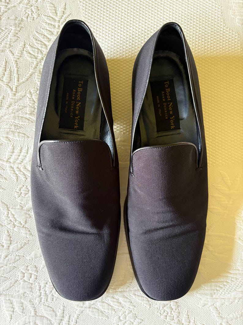 Puede incluir: Un par de zapatos sin cordones de color gris oscuro. Los zapatos tienen una puntera redondeada y un acabado liso y mate. El interior de cada zapato tiene una etiqueta que dice "To Boot New York Adam Derrick MADE IN ITALY". Los zapatos se muestran sobre una superficie blanca estampada.