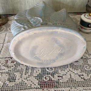 以下が含まれることがあります： 葉の模様が入った水色の陶器の花瓶。花瓶の楕円形の底には「Gonder」と「USA」の文字がエンボス加工されています。花瓶には番号「504」が記されています。