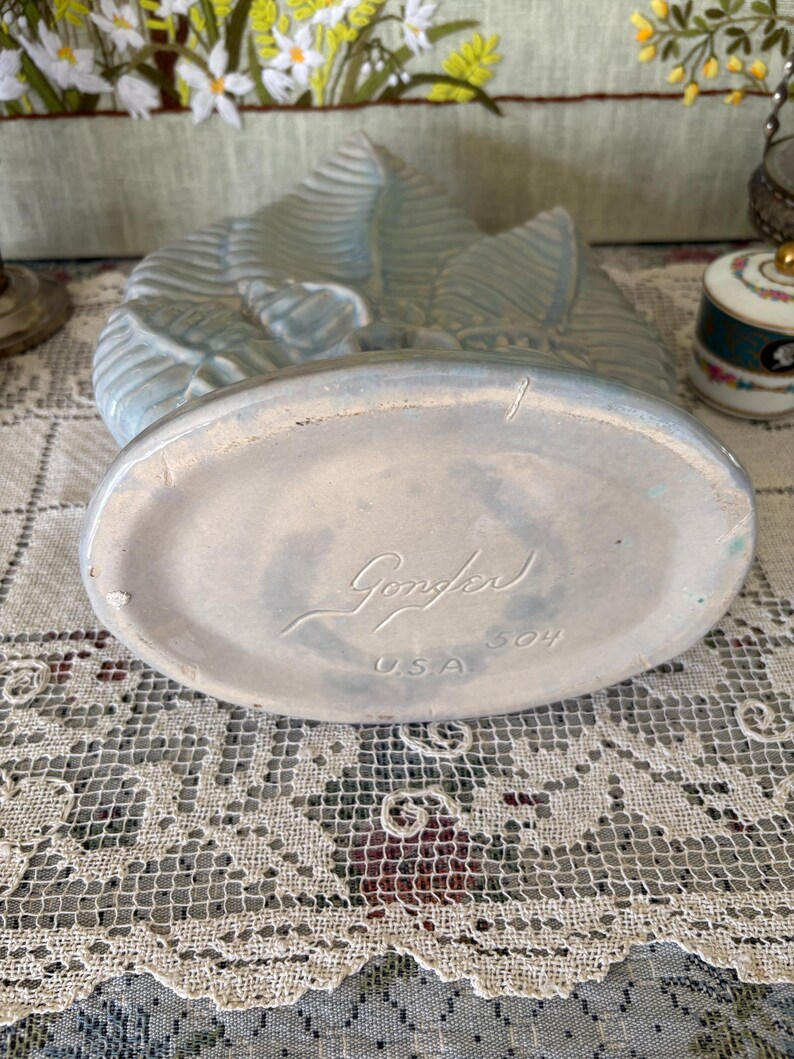 以下が含まれることがあります： 葉の模様が入った水色の陶器の花瓶。花瓶の楕円形の底には「Gonder」と「USA」の文字がエンボス加工されています。花瓶には番号「504」が記されています。