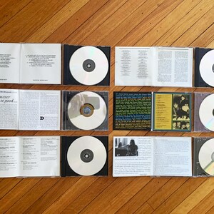 May include: A collection of eight compact discs in black jewel cases. The discs are arranged in two rows of four. The discs are all white with a silver center. The cases are all black with a silver spine. The cases are arranged on a wooden surface.