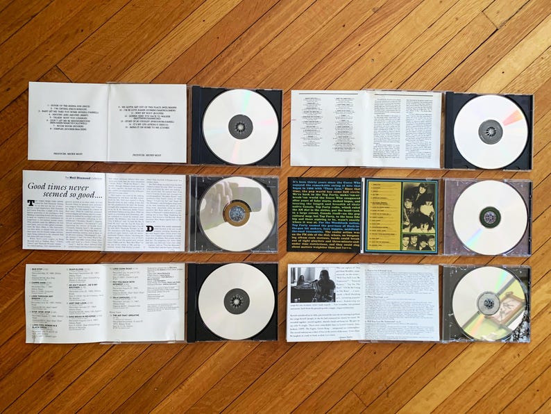 May include: A collection of eight compact discs in black jewel cases. The discs are arranged in two rows of four. The discs are all white with a silver center. The cases are all black with a silver spine. The cases are arranged on a wooden surface.