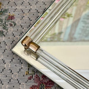 May include: A rectangular mirror with a clear glass frame featuring vertical, ribbed detailing. The corners have gold-toned metal accents. The mirror reflects a soft, blurred image, suggesting a decorative piece for home decor.