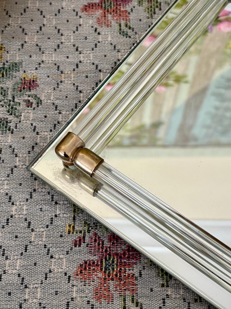 May include: A rectangular mirror with a clear glass frame featuring vertical, ribbed detailing. The corners have gold-toned metal accents. The mirror reflects a soft, blurred image, suggesting a decorative piece for home decor.