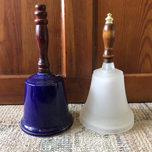 May include: Two decorative bells with wooden handles. The bell on the left is blue and white with a gold trim. The bell on the right is white with a frosted finish.