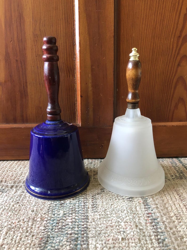 May include: Two decorative bells with wooden handles. The bell on the left is blue and white with a gold trim. The bell on the right is white with a frosted finish.