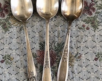 May include: Three silver teaspoons with ornate floral designs on the handles. The spoons are arranged in a row on a patterned fabric background.